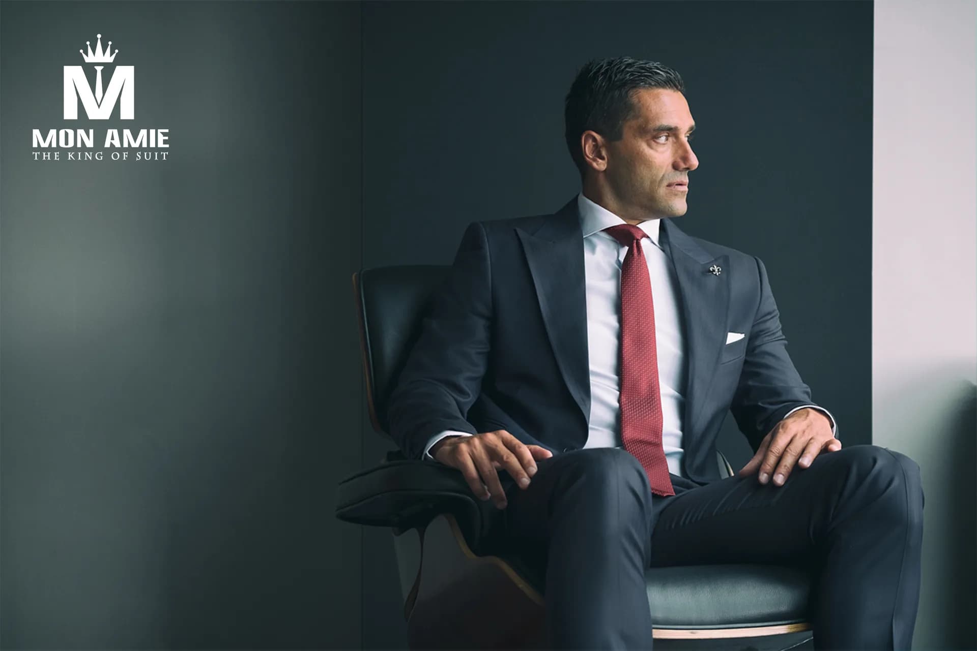 Doanh nhân mặc suit xanh navy, cà vạt đỏ, ngồi ghế da trên nền tường xám — chuẩn dress code business formal