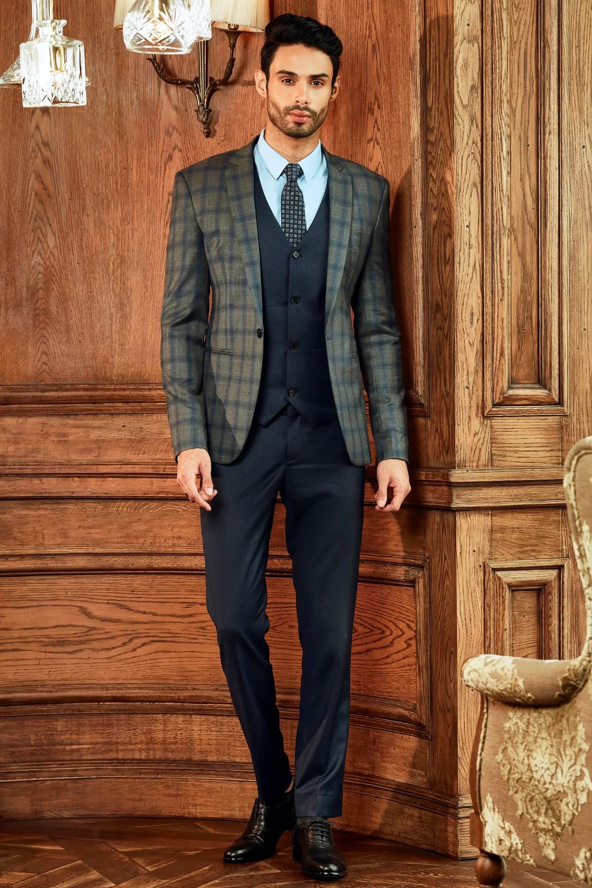 Blazer ca-rô nâu xanh, gile navy và cà vạt họa tiết trên nền ván gỗ ấm, nút cà vạt gọn gàng theo kiểu Half Windsor