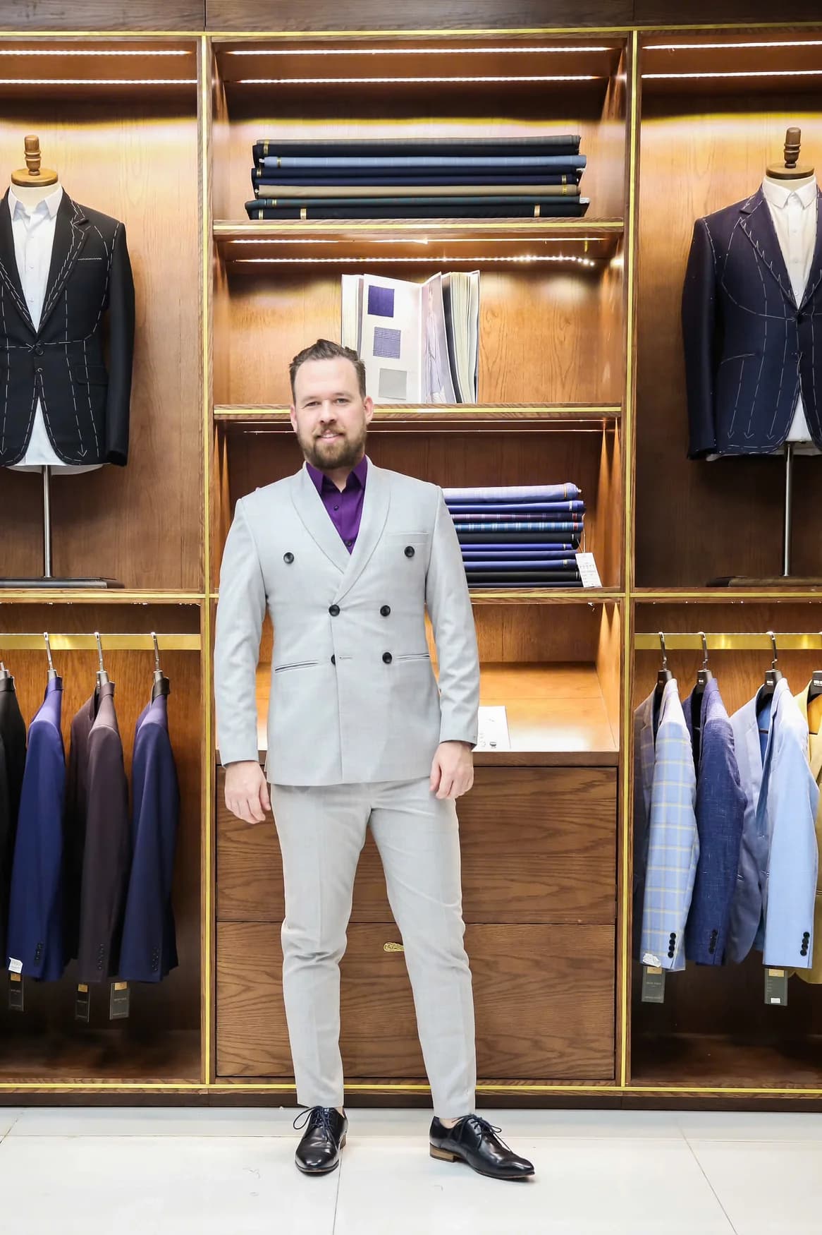 Quý ông Tây vest xám sáng, sơ mi tím, đứng trong showroom gỗ, bộ vest giữ phom tốt nhờ được chăm sóc đúng cách