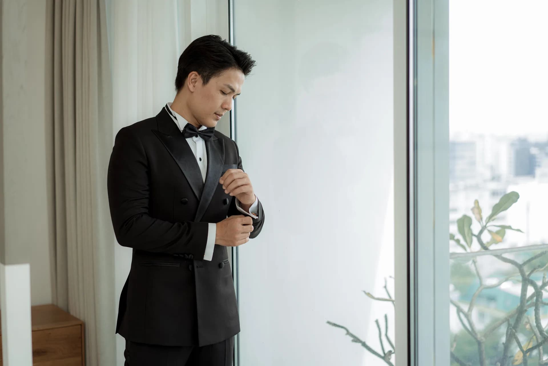 Quý ông chỉnh măng sét tuxedo bên cửa kính, dáng đứng thanh lịch, gợi ý may tuxedo đo riêng