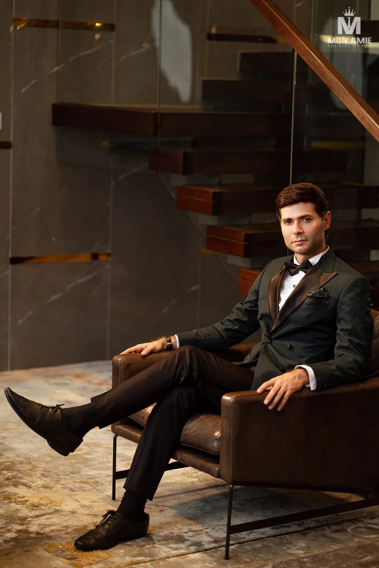 Tuxedo xanh rêu hai hàng khuy, ve satin, nơ đen, ngồi ghế da sang trọng, gợi không gian showroom Mon Amie ở khu trung tâm Sài Gòn