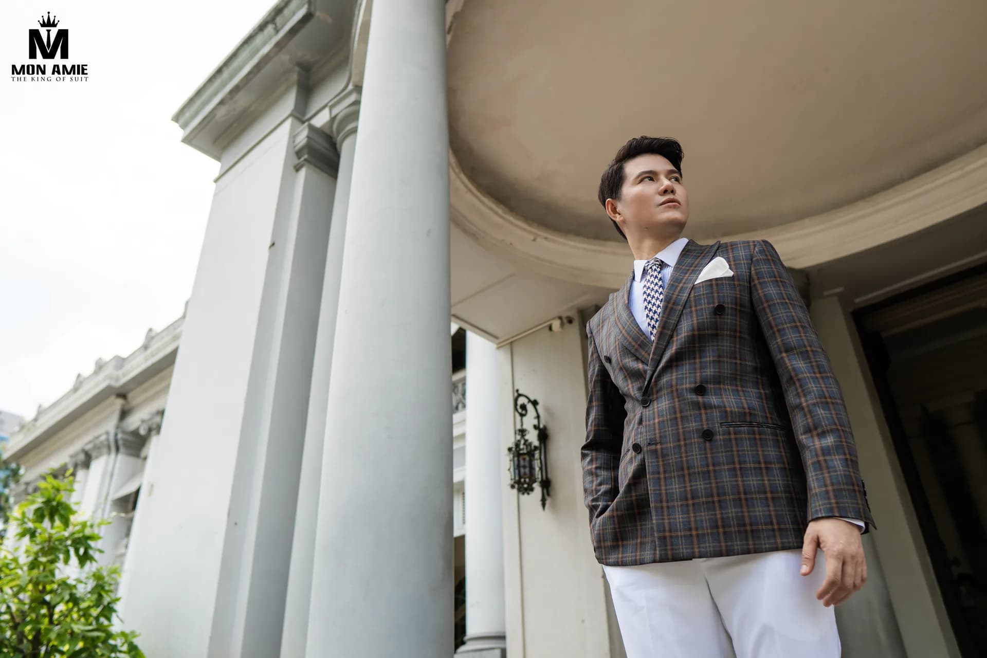 Quý ông mặc suit caro hai hàng khuy chụp góc thấp dưới hàng cột đá của toà nhà cổ, gợi ý quà tặng sếp dịp Tết bằng đồ may đo