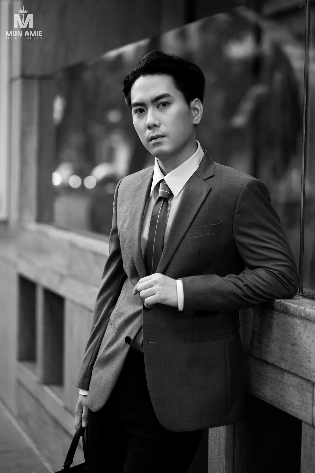 Ảnh đen trắng quý ông tựa tường kính, tay cài khuy áo vest, minh họa kiểu khuy áo vest