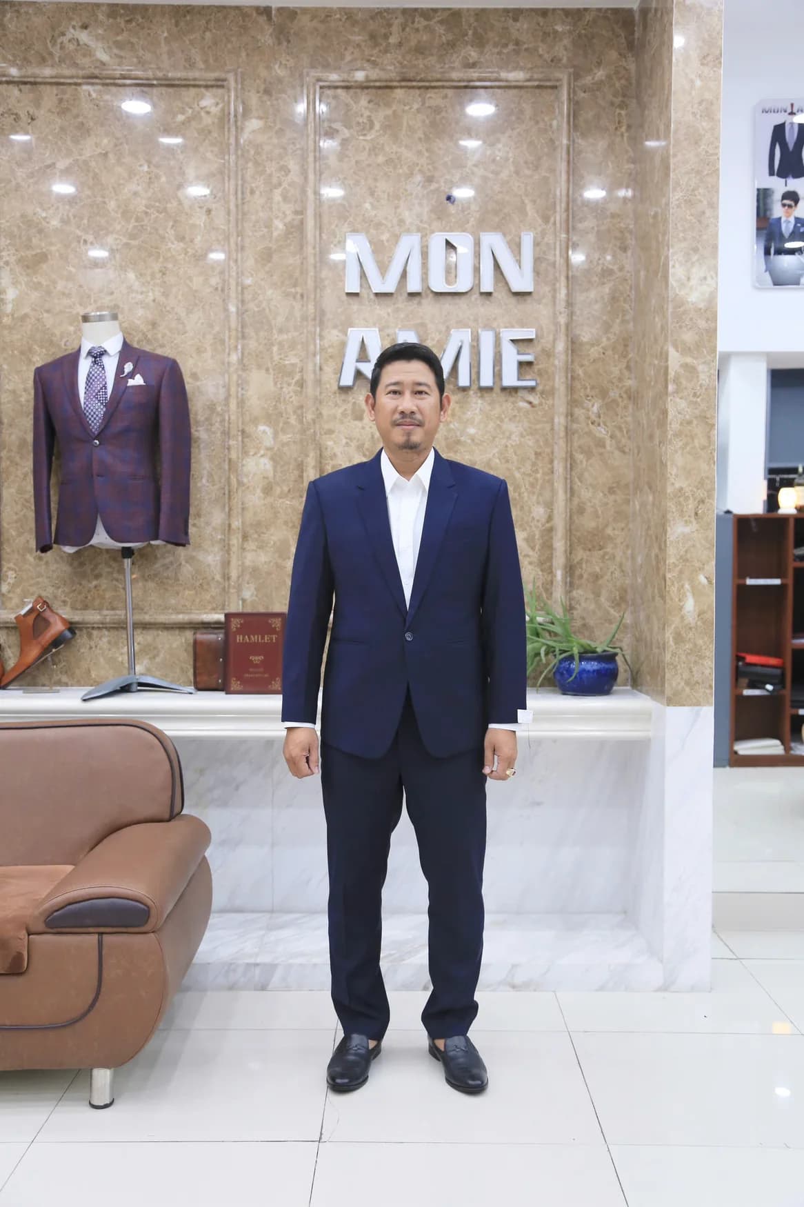 Người đàn ông mặc vest navy, sơ mi trắng đứng trước bảng hiệu Mon Amie, gợi ý quà cho người mặc vest mỗi ngày