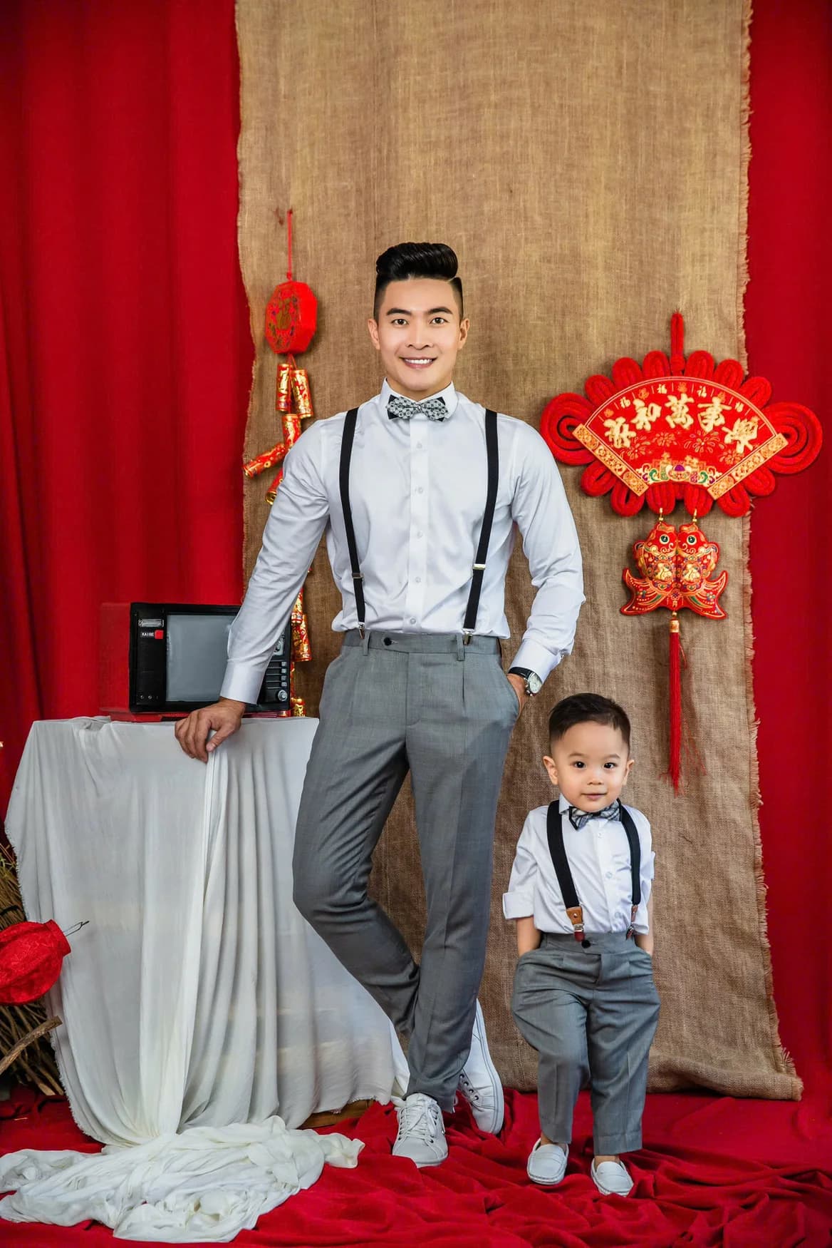 Bố và con trai mặc sơ mi trắng, quần xám dây đeo trước phông Tết đỏ, ý tưởng may áo vest nam cho bố