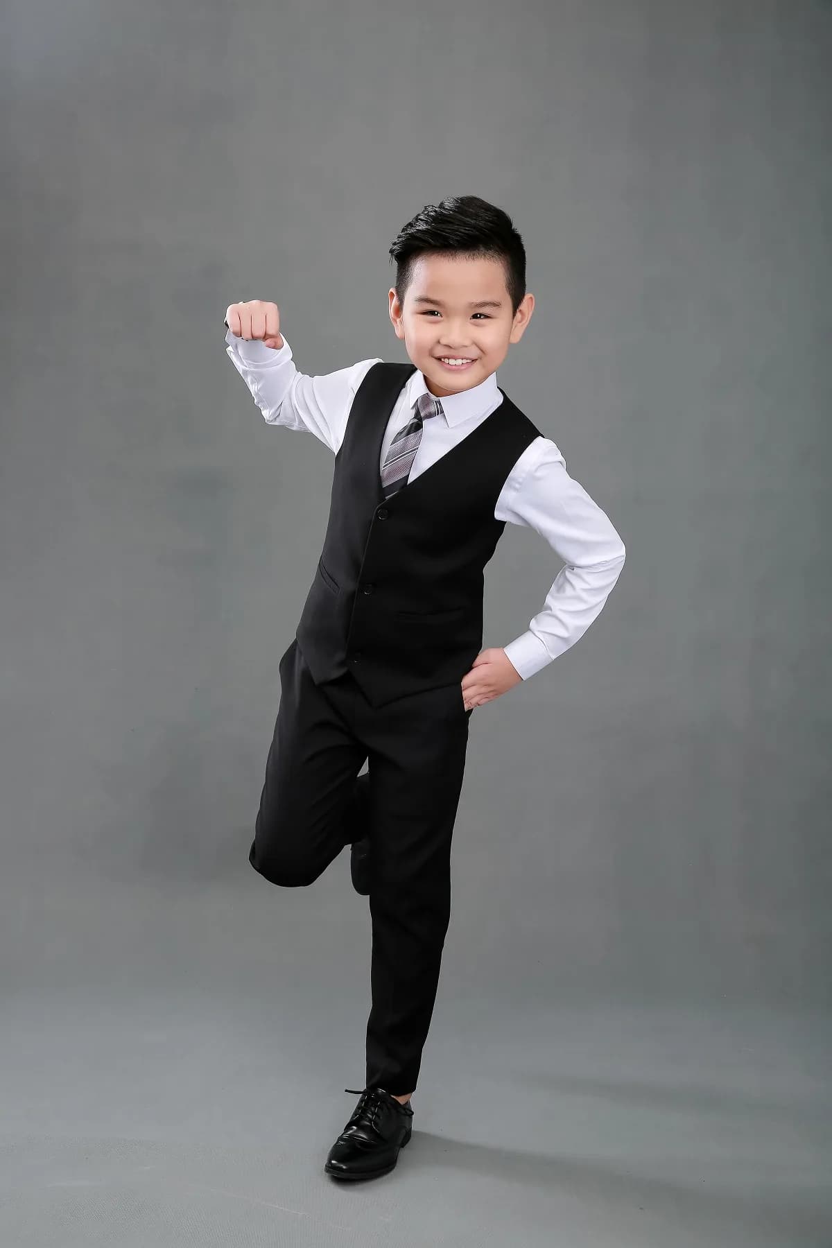 Bé tinh nghịch giơ nắm tay, cười toe toét trong bộ gile đen, minh hoạ set quà tặng đối tác cuối năm có phần dành cho bé
